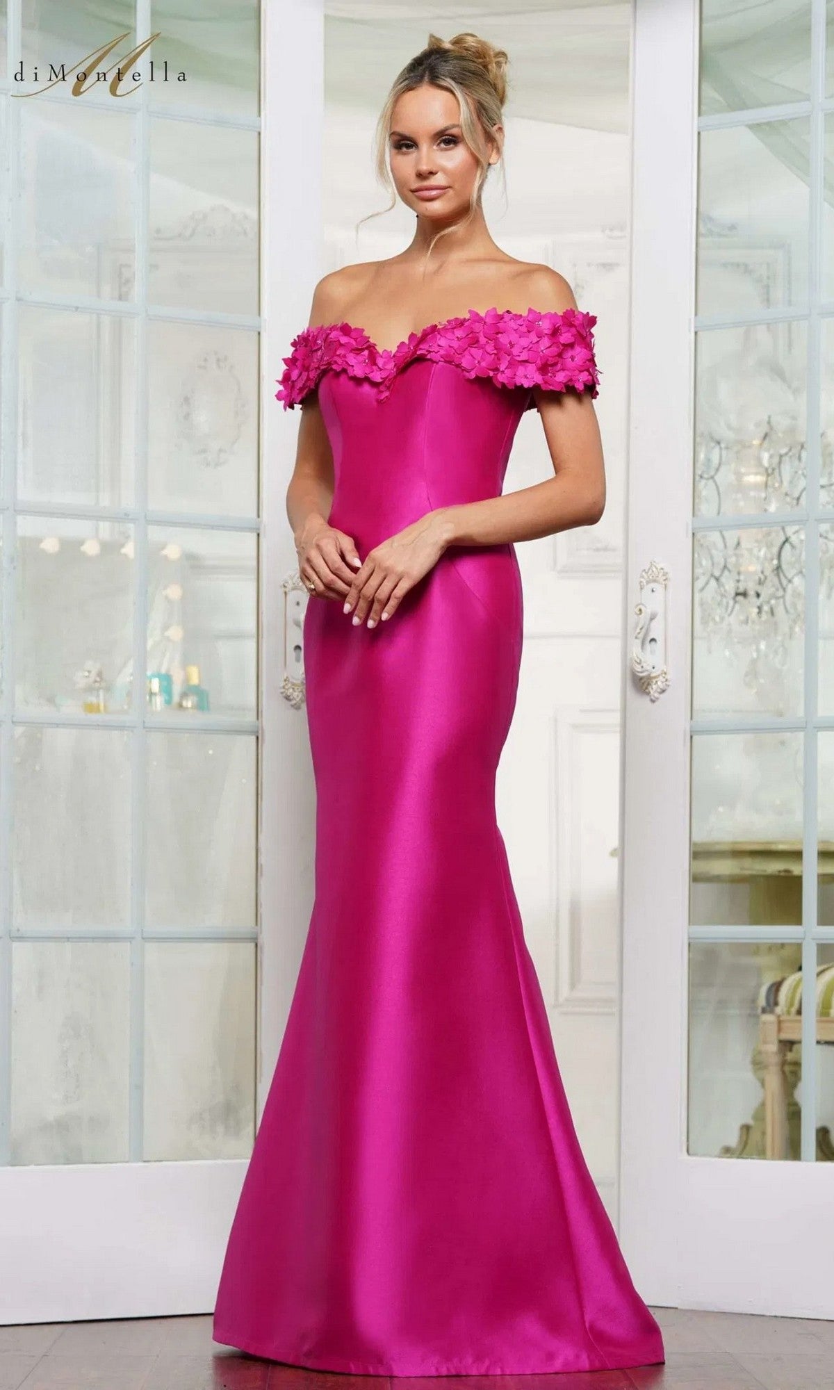 A woman stands before glass doors in the Rina Di Montella RD3254 long formal dress, a magenta off-the-shoulder gown featuring floral neckline detailing and elegant silk-like appeal.